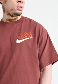 Brūns kokvilnas T-kreklis ar īsām piedurknēm, uz kura krūšu daļas atrodas sarkans un oranžs grafiskais logo, kā arī balts svītra. Gluda tekstūra.