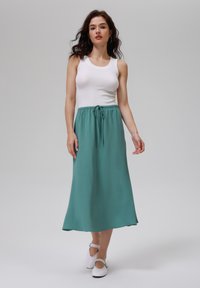 Lyse teal maxi-nederdel med en tekstureret overflade, der har en elastisk talje og snorelukning. Kombineret med en tætsiddende hvid top og hvide sko.