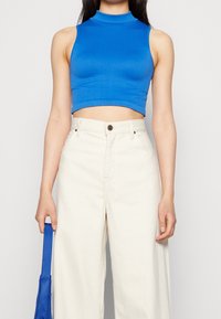 Blå ribbade crop-top med hög halsringning, i kombination med krämfärgade vida byxor med hög midja. Personen håller en blå väska i vänster hand.