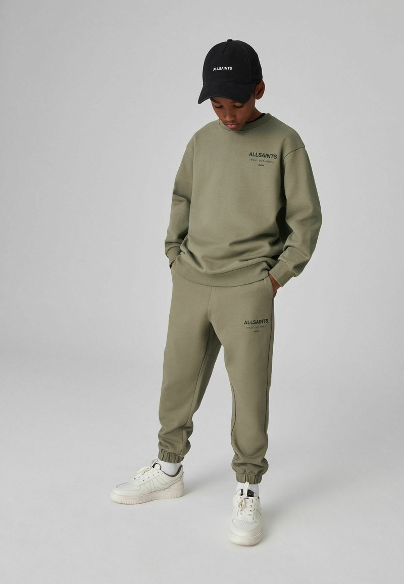 Maslinasto zeleni duksevi i trenerke s crnim logom, upareni s bijelim tenisicama. Outfit ima opušten kroj i rebraste manžete.