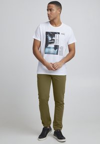 Weißes Baumwoll-T-Shirt mit Grafik eines Palme und Text, kombiniert mit olivgrünen, taillierten Hosen und schwarzen Sneakers mit weißen Akzenten.