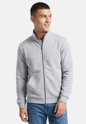 Junger Mann lächelt, trägt eine hellgraue Jacke mit Reißverschluss und Seitentaschen sowie blaue Jeans, schaut leicht nach rechts.