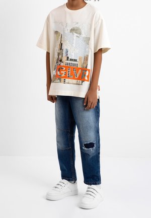 Beige t-shirt med grafiktryk og orange "GLVR"-tekst, kombineret med blå slidte denimjeans og hvide sneakers.