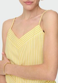 Geel gestreepte top met dunne bandjes, met een v-hals ontwerp en een lichte textuur. Een gouden hexagonale armband is te zien om de pols.