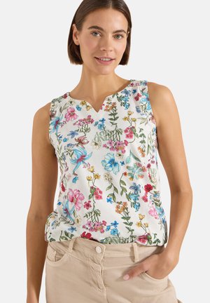 Femme portant un haut blanc sans manches avec un imprimé floral coloré et un pantalon beige, souriante avec une main dans la poche.