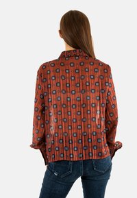 Blusa roja con patrón y diseños geométricos oscuros azules irregulares. Cuenta con un collar, mangas largas y una tela ligera y texturizada.