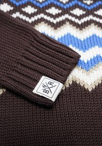 Stickad tröja i mörkbrun med texturerad ribbad manchett. Har ett geometriskt mönster i blått, vitt och beige. Inkluderar en etikett.