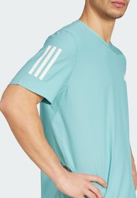 Una camiseta atlética de manga corta en color turquesa, con una tela texturizada, tres rayas blancas en el hombro izquierdo y un corte relajado.