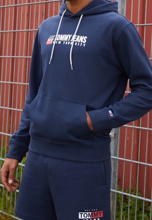 Person trägt einen dunkelblauen Tommy Jeans Hoodie und passende Shorts und steht mit einer Hand in der Tasche vor einem Metallzaun.