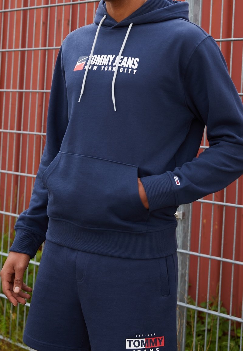 Personne portant un sweat à capuche Tommy Jeans bleu marine et un short assorti, debout avec une main dans la poche devant une clôture métallique.