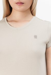 Beige fitted t-shirt with a round neck and short sleeves. Features a small embossed logo on the left chest. Smooth texture.