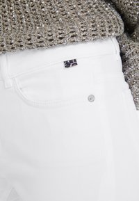 Weiße Jeans mit einer taillierten Passform, die ein kleines Leopardenmuster-Label und silberne Knopfakzente aufweisen. Der Stoff ist glatt und leicht.