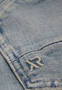 Lichtblauwe denimstof met een close-up van een zak, voorzien van een subtiele crèmekleurige geborduurde "R"-ontwerp en zichtbare stikdetails.