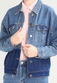 Denim jacket with a light and dark wash, large chest pockets, and metal buttons. Features a classic collar and a loose fit design.