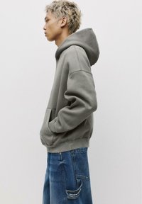 Gray fleece hoodie with a front pocket, features a drawstring hood, relaxed fit, and ribbed cuffs; worn with blue denim cargo pants.