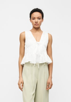 Γυναίκα που φοράει αμάνικο άσπρο τοπ πεπλούμ με υφή και δέσιμο μπροστά, συνδυασμένο με ψηλόμεσο μπεζ παντελόνι, στέκεται μπροστά σε απλό φόντο.