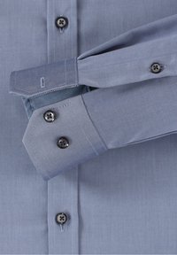 Chemise bleu clair avec une texture lisse, devant à boutons, tissu intérieur des poignets contrastant et boutons foncés. Les poignets présentent un design angulaire.