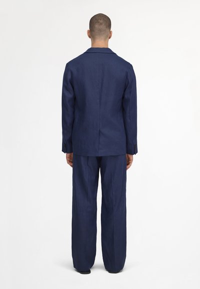 Homme debout, dos à la caméra, portant un costume bleu marine avec un pantalon ample et une veste à manches longues, sur un fond blanc.