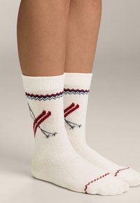 Pletene bijele čarape s uzorkom crvenih i sivih skijaških štapova, rebrasti rub i kontrastni obrub oko vrha. Meka tekstura.