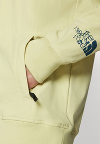 Bluza de culoare galben deschis cu un buzunar lateral și un logo bleumarin pe mânecă. Textura moale și manșetele cu nervuri oferă un confort sporit.