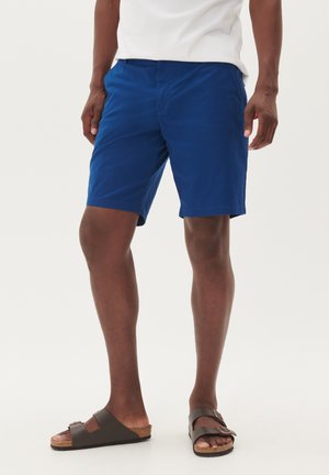 Men's blue shorts made of lightweight fabric, featuring a straight cut, side pockets, and a flat front design. Worn with brown slide sandals.