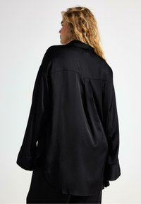 Kvinde med bølget blont hår iført en løs sort satin skjorte med langeærmer, set bagfra mod en ensfarvet hvid baggrund.