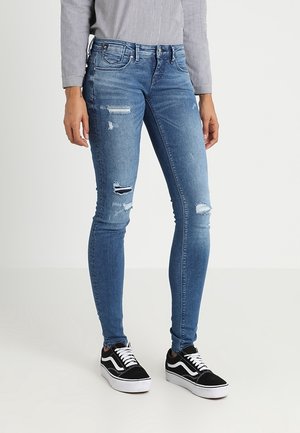 Jeans ajustados de mezclilla azul claro con detalles desgastados que incluyen roturas. Combinados con zapatillas negras y blancas. Textura suave, diseño ajustado.