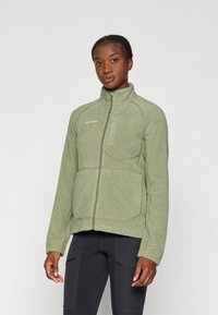 Grüne Fleecejacke mit hohem Kragen, Reißverschluss und zwei Fronttaschen. Verfügt über einen strukturierten Stoff und sichtbare Nähte.