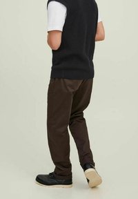 Dunkelbraune Hose mit lockerer Passform, kombiniert mit einem schwarzen Strickwesten über einem weißen T-Shirt. Die Fußbekleidung besteht aus schwarzen Schuhen mit hellen Sohlen.