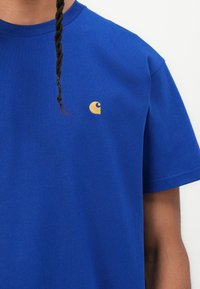 Royal blue cotton t-shirt with a round neckline and short sleeves. Features a small golden logo on the left chest. Smooth texture.
