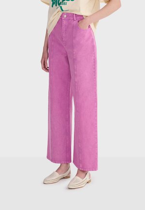 Personne portant un jean rose taille haute à jambes larges, assorti à des chaussures tissées crème sans lacets, debout avec une main dans la poche.