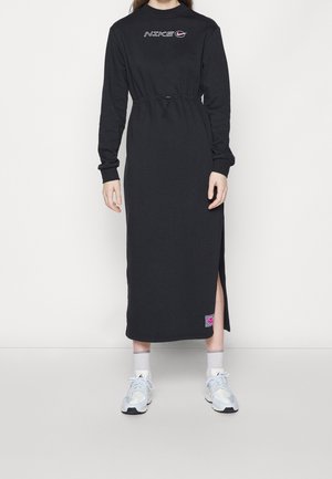 Zwarte jurk met lange mouwen en een trekkoord in de taille, met een logo op de borst en een zijsplit. Gecombineerd met lichtblauwe sportschoenen.