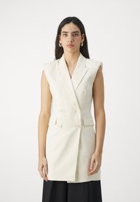 Blazer sans manches crème avec un design à double boutonnage, des revers crantés et des détails aux épaules ; il comporte deux poches avant et une texture de tissu.