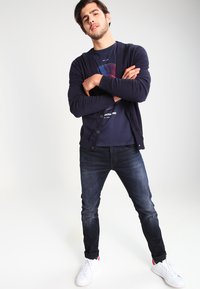 Marineblauwe cardigan over een marineblauw t-shirt met grafische print, gecombineerd met donkere spijkerbroeken en witte sneakers, wat zorgt voor een casual look.