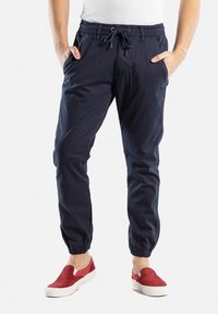 Pantalones jogger de algodón navy con puños elásticos, cintura con cordón y bolsillos laterales. Combinados con zapatillas rojas sin cordones.