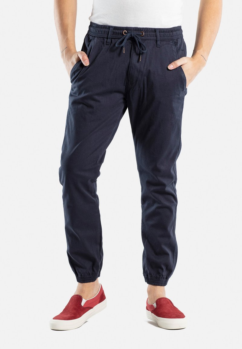 Pantalones jogger de algodón navy con puños elásticos, cintura con cordón y bolsillos laterales. Combinados con zapatillas rojas sin cordones.