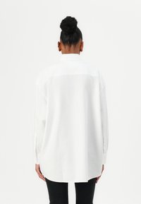Hvit oversized skjorte med lange ermer, bakryggdetalj og rund hem. Kombinert med svart bukse for kontrast.