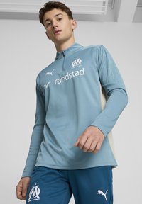 Maglietta sportiva a maniche lunghe blu chiaro con mezza cerniera, con un logo sul petto e maniche con motivo. Abbinata a pantaloni corti blu scuro.
