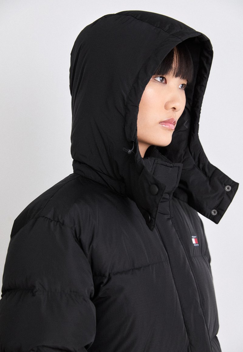 Schwarze Pufferjacke mit Kapuze, hohen Kragen, Druckknöpfen und kleinem Logo-Emblem auf der Brust. Glatte Textur und gepolstertes Design.