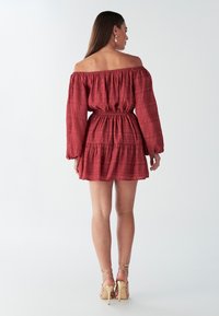 Rode off-shoulder jurk van gestructureerde stof, met een elastische tailleband, lange mouwen en een gelaagde rok. Gouden sandalen met bandjes.