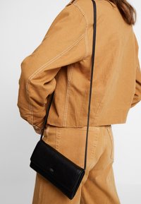 Donna che indossa giacca e pantaloni color cammello con una borsa a tracolla in pelle nera appesa alla spalla.