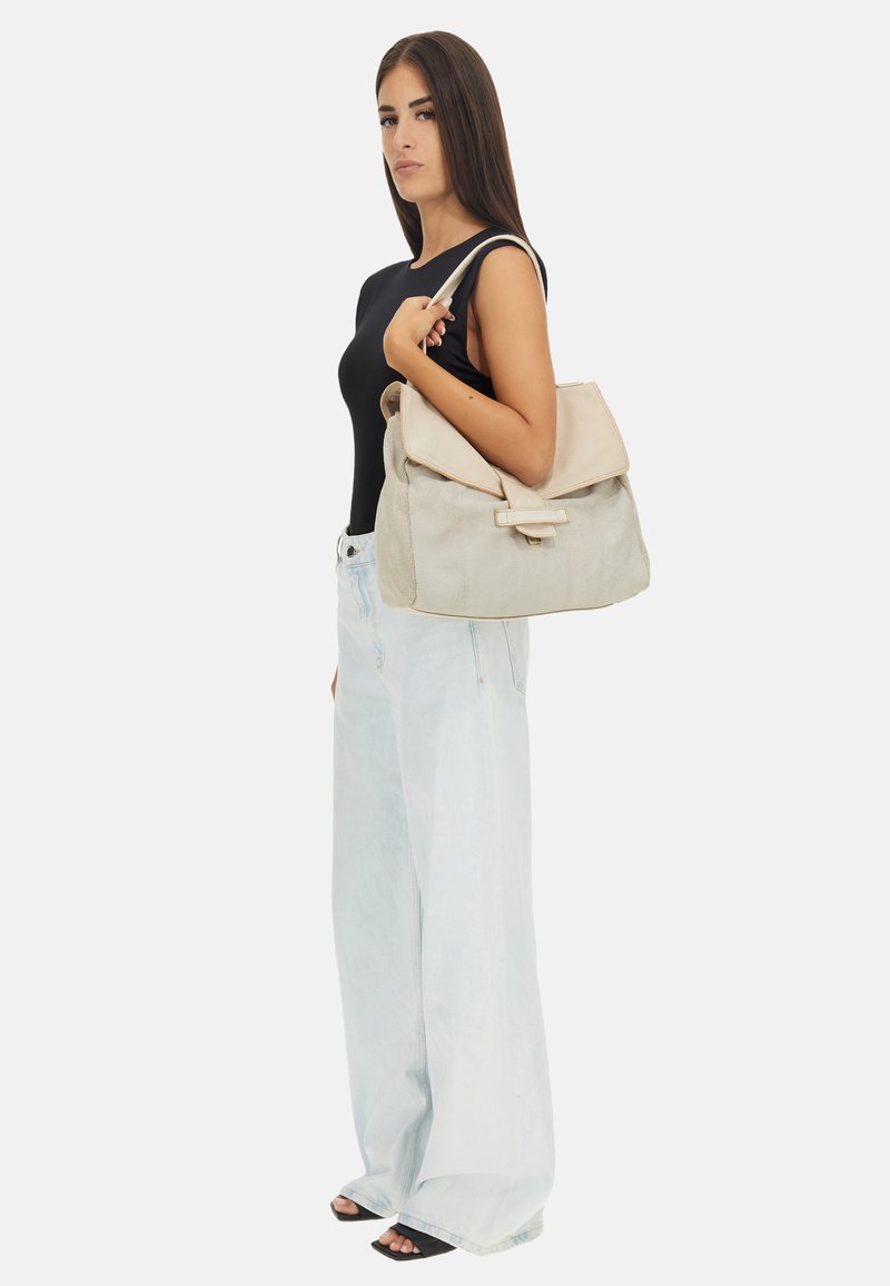 Borsa beige con texture, dotata di un flap e una fibbia frontale, portata su una spalla. Il modello indossa una maglietta nera e jeans a gamba larga di colore azzurro chiaro.