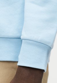 Μανσέτα φούτερ ανοιχτού μπλε με ριμπ υφή, σε συνδυασμό με σκούρο χέρι και χακί ύφασμα στο φόντο. Απαλός, καθημερινός σχεδιασμός.