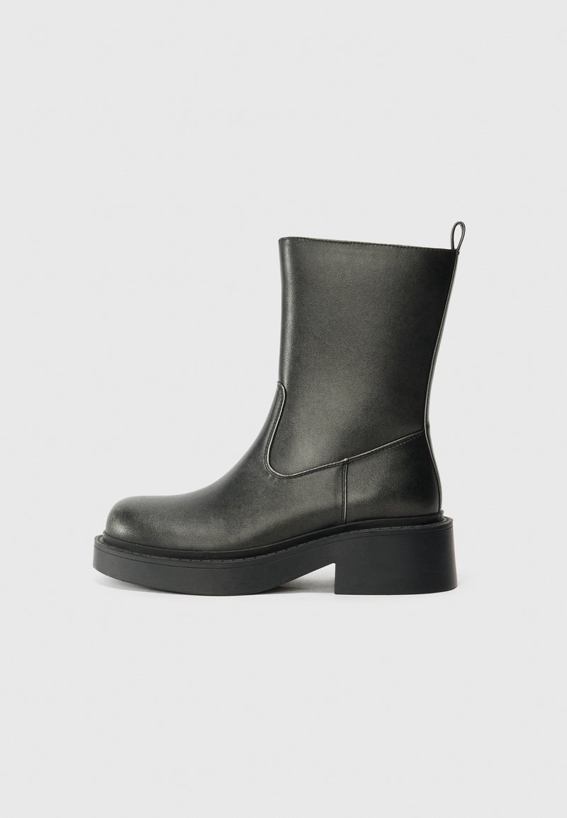 Botte en cuir noire mi-mollet avec bout arrondi, semelle épaisse, talon bloc et languette à l'arrière sur fond gris uni.
