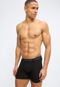 Μυώδης άνδρας με μαύρα εσώρουχα Tommy Hilfiger, κοιτάζει προς τα δεξιά, στέκεται μπροστά σε απλό ανοιχτό φόντο.