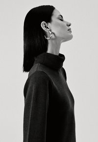 Maglione a collo alto nero con colletto a costine, texture liscia e maniche aderenti. Grandi orecchini a cerchio metallici accentuano il profilo.