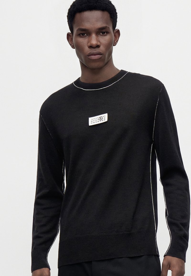 Joven con una camiseta negra de manga larga con costuras blancas y un pequeño parche rectangular en el pecho.