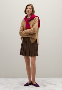 Beige Strickpullover mit gerippten Details, roter und pinker gestreifter Schal, plissierter brauner Karorock und lila Slipper.