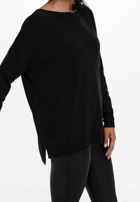 Czarny, oversize sweter z prążkowanymi mankietami i dołem, z wysokim kołnierzem oraz bocznymi rozporkami. Noszony z czarnymi legginsami. Miękka tekstura.