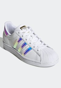 White leather sneaker with a rounded toe, three iridescent stripes, and a textured rubber sole. Features a gold logo on the tongue.